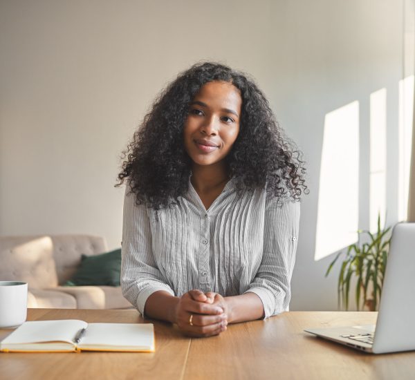 Portrait of positive self confident young dark skinned female teacher with voluminous hairdo getting ready for online lesson, sitting at desk with laptop, coffee and copybook in home office interior